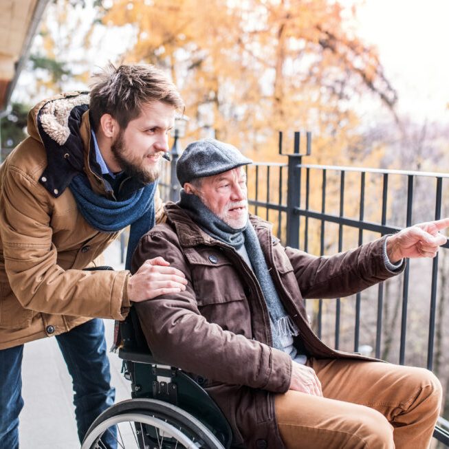 Father in wheelchair and young son on a walk. A carer assisting disabled senior man.